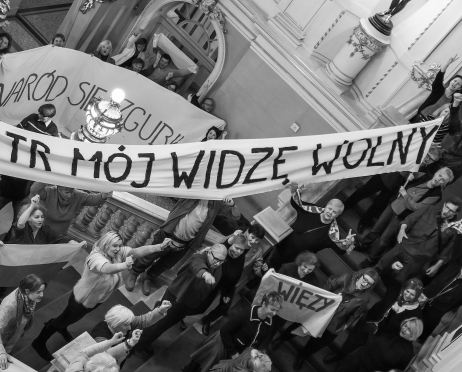 Czarno-białe zdjęcie. Grupa protestujących zgromadzonych na ozdobnej klatce schodowej we wnętrzu teatru. W przestrzeni pełnej stiuków i balustrad rozwieszone są duże transparenty z hasłami „Teatr mój widzę, wolny” oraz „Naród się zgubił”. Wiele osób unosi pięści i ręce w geście sprzeciwu.